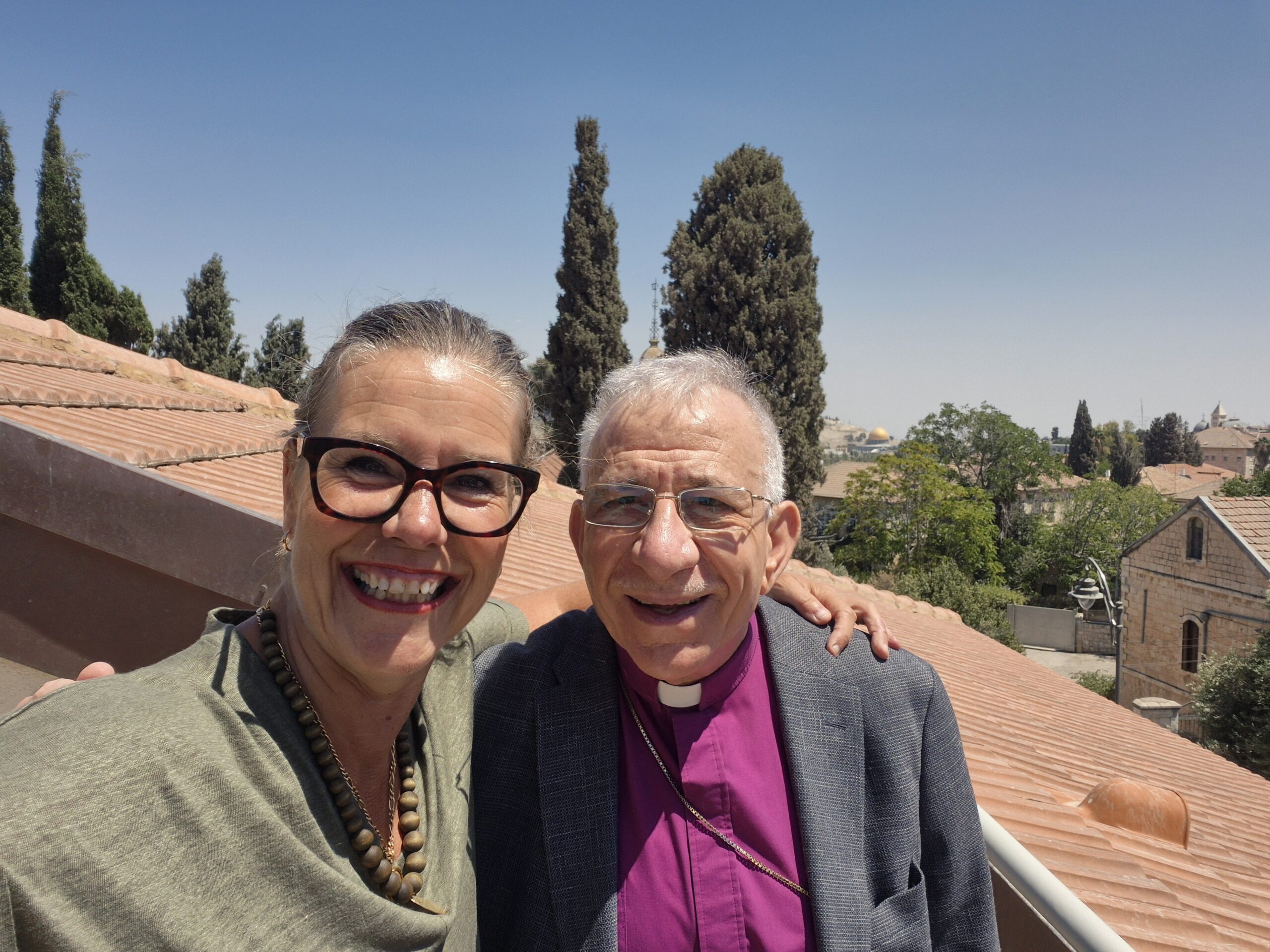 Nainen ja mies seisovat vierekkäin hymyillen. Taustalla näkyy punatiilinen katto ja sen takana Jerusalemin maisemia.