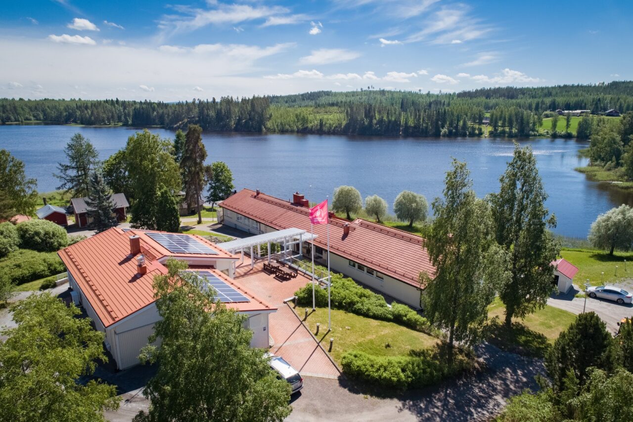 Droonikuva, jossa näkyy U:n muotoinen rakennus, metsää ja järveä kesällä.
